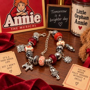 Könnte beinhalten: Ein silberfarbenes Bettelarmband mit verschiedenen Perlen in Rot, Weiß und Metallic-Silber sowie hängenden Anhängern in Form eines Hundes, eines Mädchens, einer Sonne und eines New York-Schildes an einer Gliederkette mit Karabinerverschluss.