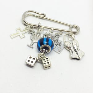 Puede incluir: Un broche de seguridad plateado con varios abalorios unidos, incluyendo una cruz, una copa de martini, una cuenta azul y negra a rayas, un dado, una carta de juego y un corsé.