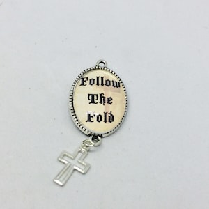 Può includere: Un ciondolo ovale argentato con il testo "Follow the Fold" in lettere nere su uno sfondo crema. Un ciondolo a croce argentato pende dalla parte inferiore del ciondolo ovale.