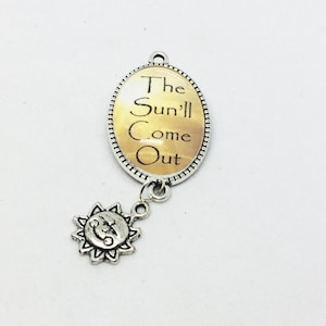 May include: An oval silver-toned pendant featuring a yellow and white gradient background with the black text The Sun'll Come Out. A small silver-toned sun charm with a smiling face is attached to the bottom of the pendant by a jump ring.