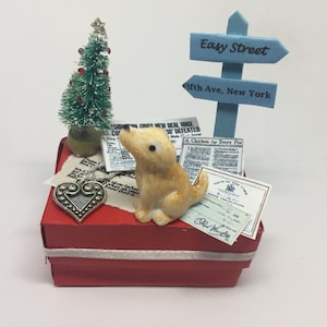 May include: A red box with a white ribbon, a small green Christmas tree, a small ceramic dog, and a sign that reads "Easy Street" and "Fifth Ave, New York".
