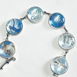 Könnte beinhalten: Ein silbernes Armband mit sechs runden Glasanhängern mit Bildern und Texten aus dem Film Frozen. Die Anhänger zeigen eine Silhouette von Elsa, den Text "Let it Go!" und "Frozen", ein Bild von Olaf, den Text "Love is a Force of Nature" und ein Bild von Sven.