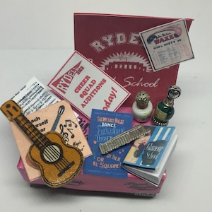 Può includere: Una scena in miniatura a tema scolastico con una scatola rosa, una piccola chitarra, un foglio di musica, un cartello che dice "Rydell High Cheer Squad Auditions Today!" e un cartello che dice "Saturday Night Dance Entertainment You're Invited! Or Treat Be Square!"