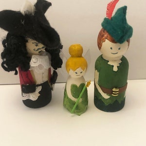 Könnte beinhalten: Drei handbemalte Holz-Steckpuppen, die Figuren aus der klassischen Geschichte Peter Pan darstellen. Die Puppen sind Captain Hook, Tinkerbell und Peter Pan. Captain Hook trägt einen schwarzen Hut, eine rote Jacke und eine schwarze Hose. Tinkerbell trägt ein grünes Kleid und hält einen grünen Zauberstab. Peter Pan trägt einen grünen Hut, ein grünes Hemd und eine braune Hose.