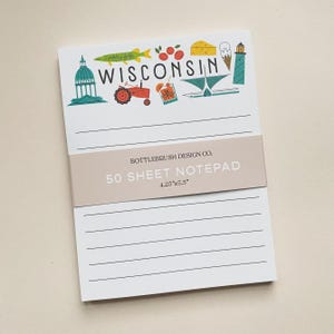 May include: A white notepad with horizontal lines and a tan band that reads "BOTTLERUSH DESIGN CO. 50 SHEET NOTEPAD 4.25"x5.5"". The top of the notepad features colorful illustrations and the word "WISCONSIN".