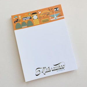 May include: White notepad with an orange header featuring illustrations of a clock tower, bridge, and other city icons. The word "Milwaukee" is written in cursive at the bottom.