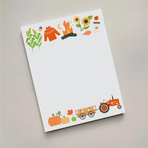 May include: A white notepad with colorful fall-themed illustrations. The top border features a sweater, campfire, sunflower, and coffee cup. The bottom border includes pumpkins, a tractor, and a wagon. The notepad is perfect for jotting down notes.