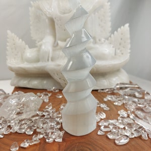 Puede incluir: Una torre de selenita blanca con un diseño en espiral, rodeada de cristales de cuarzo transparentes sobre una superficie de madera.