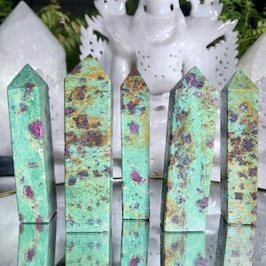 Può includere: Sei punte di cristallo di zoisite verde e rosso rubino con una punta appuntita e una base piatta. I cristalli sono disposti in fila su una superficie riflettente.