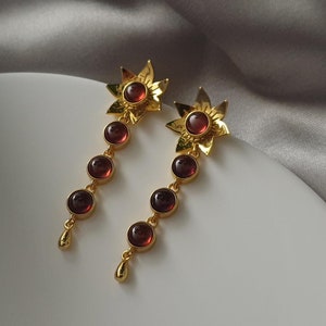 May include: A pair of gold earrings with a flower design and red gemstones. The earrings are dangling and have a drop shape.