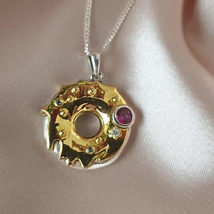 May include: Gold and silver pendant necklace with a donut-shaped design. The donut is decorated with small blue and white gemstones and a larger red gemstone.
