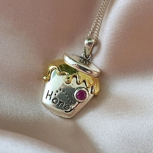 Può includere: Una collana con ciondolo a forma di barattolo di miele in argento e oro con una pietra preziosa rosa. Il ciondolo ha incisa la parola "Honey".