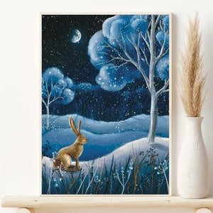 Könnte beinhalten: Gerahmter Kunstdruck, der eine Winterlandschaft mit einem braunen Hasen in einer verschneiten Landschaft unter einem sternenklaren Nachthimmel zeigt. Das Kunstwerk zeigt blau getönte Bäume und einen Halbmond und schafft eine ruhige Atmosphäre. Der Druck wird auf einem Holzregal ausgestellt.