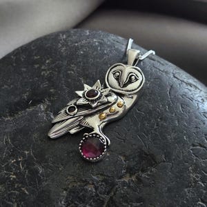 Puede incluir: Colgante de plata con forma de b&uacute;ho con un dise&ntilde;o detallado. El colgante presenta una estrella y una gema grande, redonda y de color rojo oscuro. Peque&ntilde;os detalles dorados son visibles en el cuerpo del b&uacute;ho. El colgante est&aacute; en una cadena de plata.