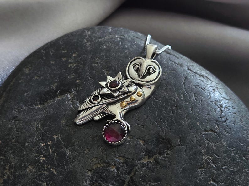 Puede incluir: Colgante de plata con forma de b&uacute;ho con un dise&ntilde;o detallado. El colgante presenta una estrella y una gema grande, redonda y de color rojo oscuro. Peque&ntilde;os detalles dorados son visibles en el cuerpo del b&uacute;ho. El colgante est&aacute; en una cadena de plata.