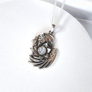 May include: Silver pendant necklace featuring a detailed design of a bird with outstretched wings holding a flower with a moonstone in the center.
