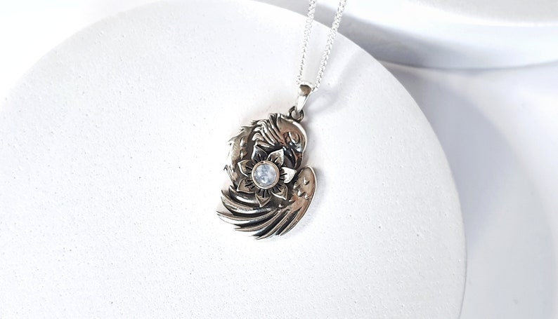May include: Silver pendant necklace featuring a detailed design of a bird with outstretched wings holding a flower with a moonstone in the center.
