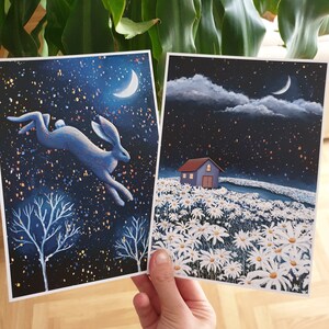 Puede incluir: Dos tarjetas ilustradas con un fondo de cielo nocturno. La primera tarjeta presenta un liebre azul saltando a trav&eacute;s de un cielo estrellado con una luna creciente. La segunda tarjeta muestra una peque&ntilde;a casa con un techo blanco en un campo de margaritas blancas bajo un cielo estrellado con una luna creciente.