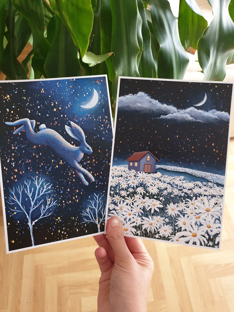 Puede incluir: Dos tarjetas ilustradas con un fondo de cielo nocturno. La primera tarjeta presenta un liebre azul saltando a trav&eacute;s de un cielo estrellado con una luna creciente. La segunda tarjeta muestra una peque&ntilde;a casa con un techo blanco en un campo de margaritas blancas bajo un cielo estrellado con una luna creciente.