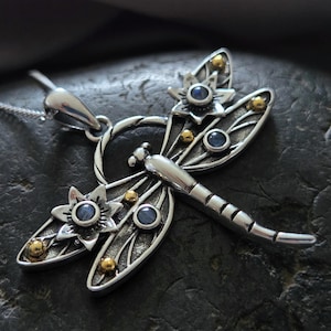 Pu&ograve; includere: Collana con ciondolo a forma di libellula d'argento con accenti di pietre preziose blu e dettagli dorati. La libellula ha ali intricate e un corpo dettagliato.