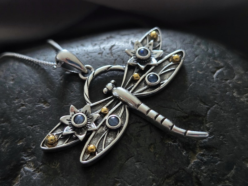 Pu&ograve; includere: Collana con ciondolo a forma di libellula d'argento con accenti di pietre preziose blu e dettagli dorati. La libellula ha ali intricate e un corpo dettagliato.