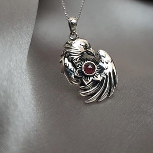 May include: Silver pendant necklace featuring a detailed design of two birds intertwined with a red gemstone in the center.
