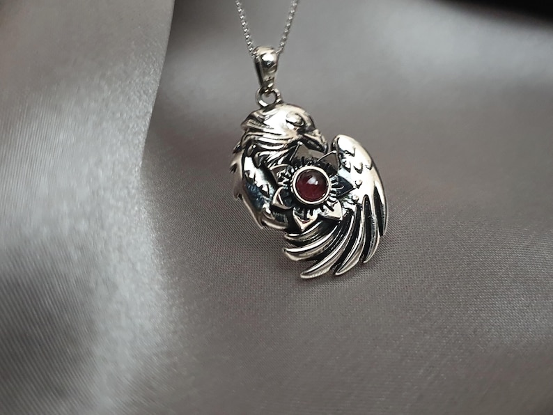 May include: Silver pendant necklace featuring a detailed design of two birds intertwined with a red gemstone in the center.