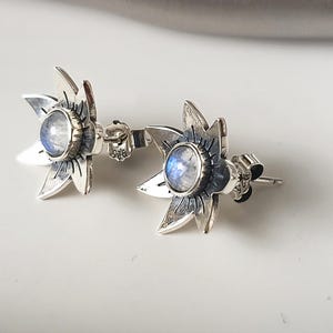 May include: A pair of silver earrings with a moonstone flower design. Each earring features a round moonstone set in a silver flower with five petals.