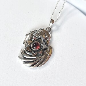 May include: Silver pendant necklace featuring a detailed design of a bird with wings spread, holding a red gemstone flower in its talons.