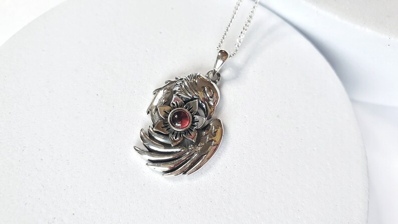 May include: Silver pendant necklace featuring a detailed design of a bird with wings spread, holding a red gemstone flower in its talons.