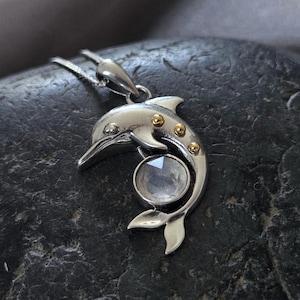 May include: Silver dolphin pendant necklace with a clear crystal gemstone and gold accents. The dolphin is holding the gemstone in its mouth.