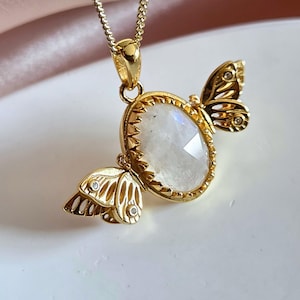 May include: Gold-toned butterfly pendant necklace. The pendant features a central oval-shaped, iridescent gemstone, framed by a scalloped gold setting. Butterfly wings are on either side, with small clear stones. The necklace chain is also gold.