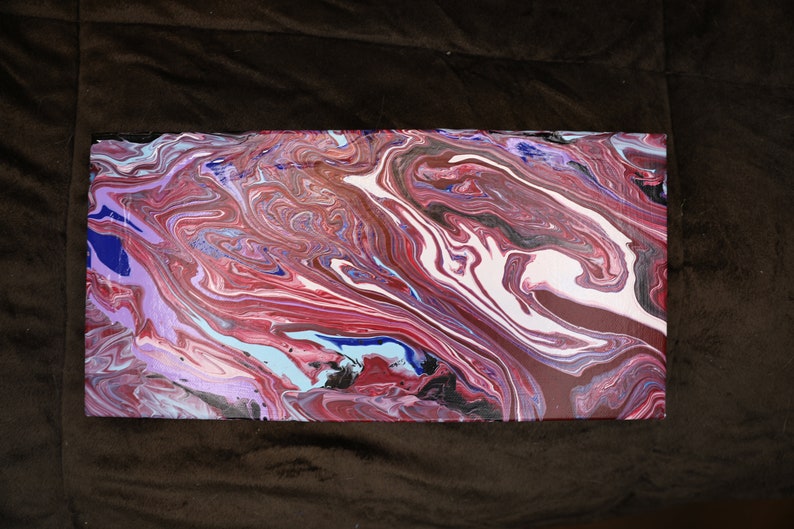May include: Rectangular abstract painting with a fluid, marbled design. The artwork features a blend of colours, including shades of red, white, blue, and black, creating a dynamic and swirling pattern. The canvas is rectangular.
