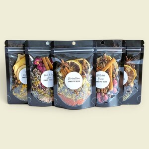May include: Five black stand-up pouches containing dried botanical potpourri blends. Each pouch features a clear front window, a circular white label with text including Love & Self Love, Divination, and Witches Brew, and visible dried fruit, cinnamon sticks, and herbs.