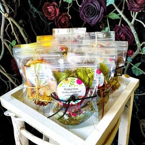 May include: A collection of clear plastic pouches filled with dried botanicals, spices, and herbs. Each pouch features a round label with text including Protection Intention Simmer Pot and Earth Magic Apothecary, tied with a black cord.