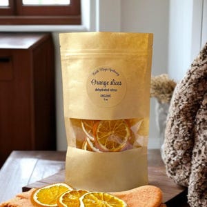 May include: A kraft paper bag with a clear window, filled with dehydrated orange slices. The label reads "Orange slices, dehydrated citrus, ORGANIC, 1 oz." Three orange slices rest on an orange cloth in front of the bag.