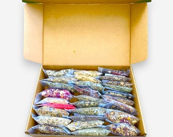 May include: An open cardboard box containing numerous small, clear plastic bags filled with various dried herbs, flowers, and botanical ingredients. The contents display a range of colors including green, brown, yellow, pink, and red.