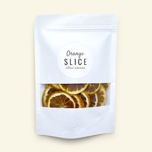 May include: A white stand-up pouch with a clear window containing dried orange slices. A circular label on the front features the text Orange SLICE citrus sinensis in black lettering. The packaging is opaque white with a transparent bottom section.