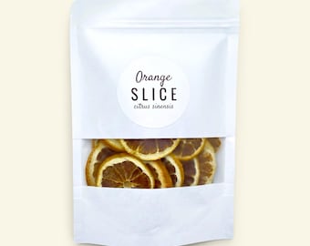 May include: A white stand-up pouch with a clear window containing dried orange slices. A circular label on the front features the text Orange SLICE citrus sinensis in black lettering. The packaging is opaque white with a transparent bottom section.
