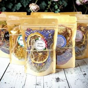 May include: Several stand-up pouches made of light brown paper with clear plastic windows, each filled with a mixture of dried herbs, flowers, and fruit slices. The front pouch features a round label with the text Home Blessings.