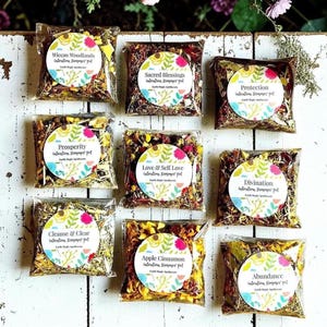 May include: Nine square clear plastic pouches filled with dried herbs, flowers, and spices. Each pouch features a round white label with colorful floral illustrations and text including Wiccan Woodlands, Sacred Blessings, Protection, Prosperity, Love & Self Love, Divination, Cleanse & Clear, Apple Cinnamon, and Abundance.