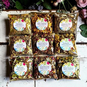 May include: Nine square clear plastic pouches filled with dried herbs, flowers, and spices. Each pouch features a circular white label with colorful floral borders and text including Abundance, Apple Cinnamon, Cleanse & Clear, Divination, Love & Self Love, Prosperity, Protection, Sacred Blessings, and Wiccan Woodlands.