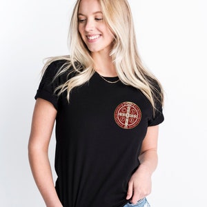 May include: A black t-shirt with a red and gold Saint Benedict medal on the left chest. The medal has the words "CSSPX" and "INRI" on it.