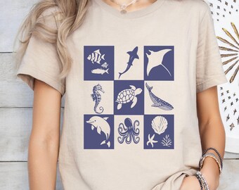 May include: Tan-colored t-shirt featuring a grid of marine life illustrations in navy blue. The design includes fish, a shark, a manta ray, a seahorse, a turtle, a whale, a dolphin, an octopus, a seashell, and coral. The word "Tan" is printed on the bottom right.