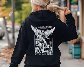May include: Black hoodie featuring a white graphic of Saint Michael the Archangel, with the text "Saint Michael the Archangel" above the image and "Defend Us In Battle" below. The hoodie is worn by a person with blonde hair.