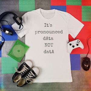 May include: A white t-shirt with the text "It's pronounced dAta NOT datA" on a background of colorful squares with binary code.