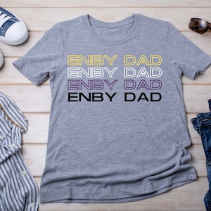 May include: A gray t-shirt with the text "ENBY DAD" repeated four times in yellow, white, purple, and black. The text is outlined in a thin, yellow line. The shirt is laid flat on a wooden surface with a pair of blue jeans, a button-down shirt, and sunglasses.