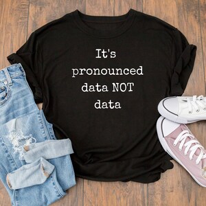 May include: Black t-shirt with white text that reads "It's pronounced data NOT data". The shirt is laid flat on a wooden surface with a pair of blue jeans and white and pink sneakers.