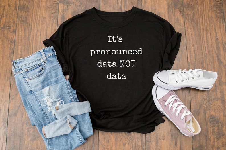 May include: Black t-shirt with white text that reads "It's pronounced data NOT data". The shirt is laid flat on a wooden surface with a pair of blue jeans and white and pink sneakers.