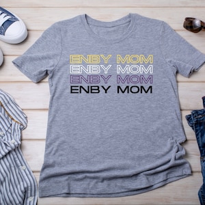 May include: Gray t-shirt with the text "ENBY MOM" repeated four times in different colors, including yellow, white, purple, and black. The shirt is laid flat on a wooden surface with a pair of blue jeans, a blue and white striped shirt, and a pair of blue sneakers.